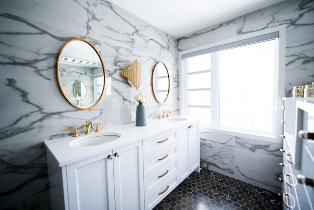 A white marble bathroom with plenty of natural light and two round mirrors.