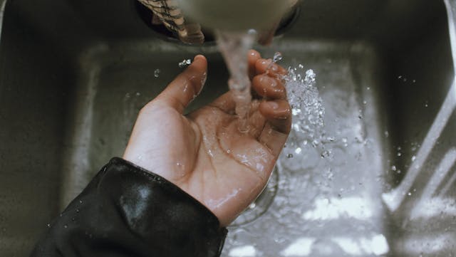 close-up of a hand under running water from a faucet in a metal sink.